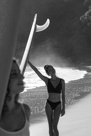 Woman in a black bikini holding a surfboard on a beach with palm trees in the background
